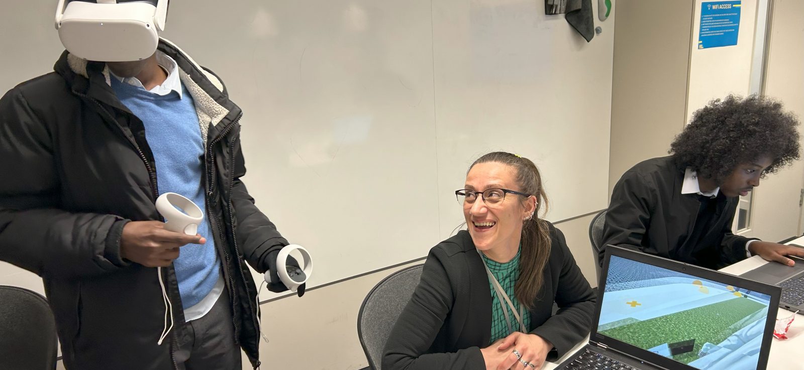 Workshop with a young male wearing a VR headset. A female teacher is smiling at him while another male pupil works at a laptop in the background.