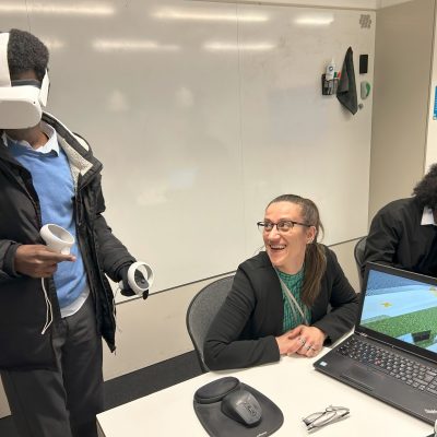 Workshop with a young male wearing a VR headset. A female teacher is smiling at him while another male pupil works at a laptop in the background.