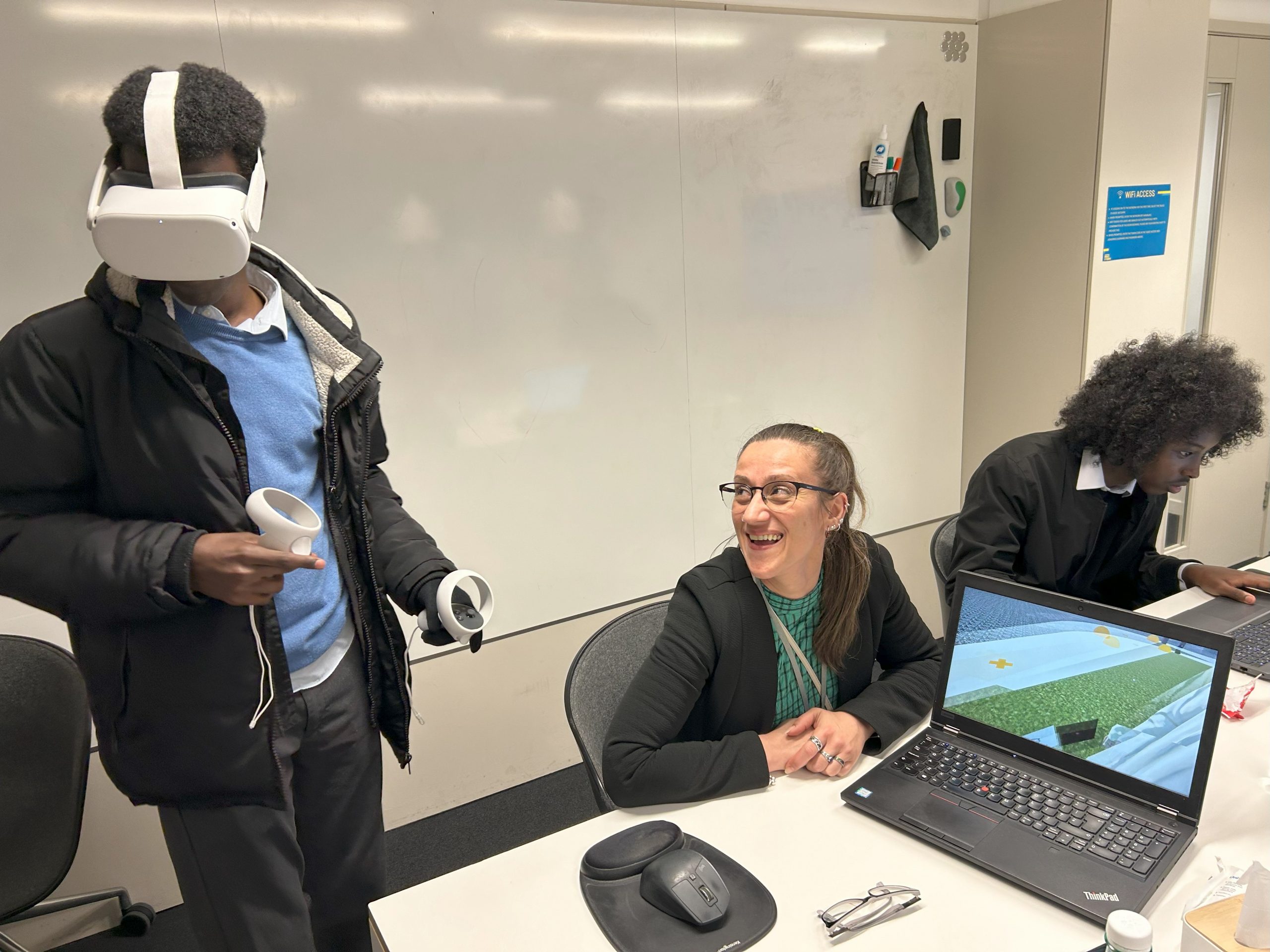 Workshop with a young male wearing a VR headset. A female teacher is smiling at him while another male pupil works at a laptop in the background.